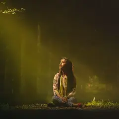 En ung kvinna sitter i skogen, badad i ett gyllene ljus. Hon ser uppåt, som om hon söker svar, kanske om vad gaia betyder.