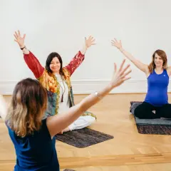 Tre kvinnor sitter på yogamattor med armarna uppåt. En av dem är gravid. En inspirerande yoga utbildning distans.