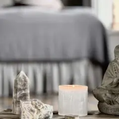En stilla Buddha-statyett, en tänd ljuslykta och stenar, kanske med honungskalcit egenskaper, på ett träbord framför en säng.