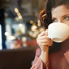 En kvinna med mörkt hår dricker kaffe ur en vit kopp. Hon har en stickad tröja på sig och sitter på ett café. Kanske är det en morgon med fastande mage kaffe.