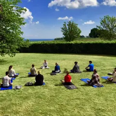 En grupp människor sitter på yogamattor på en gräsmatta under en klarblå himmel. En instruktör leder dem i en övning under ett yoga weekend.