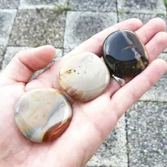 Hand håller tre polerade stenar. En mörk, en ljus med bruna strimmor, och en med blå och bruna mönster. Dessa polykrom jaspis egenskaper är fascinerande.