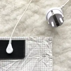 En vit jordad kabel med kontakt ligger på en vit, fluffig yta. En del av en rutig duk med en svart ficka syns. Lavasten egenskaper kan vara relaterade till jordning.