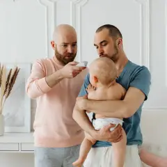 Två pappor kollar febern på sitt barn. En pappa håller barnet, den andra mäter temperaturen. Frågan "hur får man bort snuva" kan dyka upp när man tar hand om små barn.