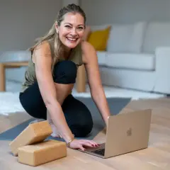 Glad kvinna förbereder sig för en lugnande restorative yoga-session med hjälp av en laptop och yogablock.