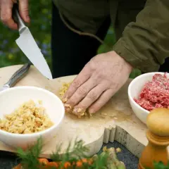 Förbereder eko mat utomhus. En hand hackar ingredienser på en träskärbräda bredvid skålar med köttfärs och en annan ingrediens.