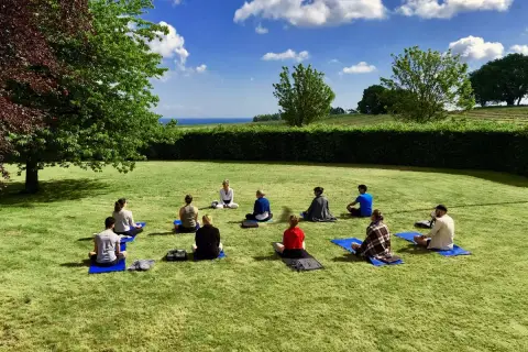 En grupp människor sitter på yogamattor på en gräsmatta under en klarblå himmel. En instruktör leder dem i en övning under ett yoga weekend.