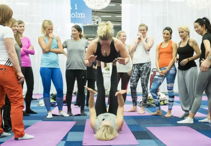 Grupp kvinnor deltar i en yogaklass. En kvinna lyfter en annan i en partnerövning, vilket visar den fysiska och andliga aspekten av yoga, liknande en andlig religion.
