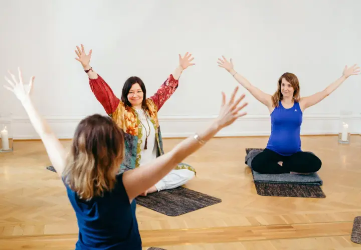 Tre kvinnor sitter på yogamattor med armarna uppåt. En av dem är gravid. En inspirerande yoga utbildning distans.