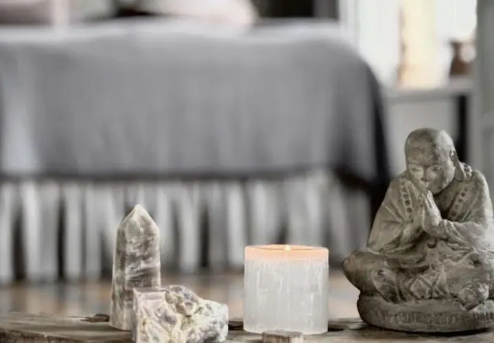 En stilla Buddha-statyett, en tänd ljuslykta och stenar, kanske med honungskalcit egenskaper, på ett träbord framför en säng.