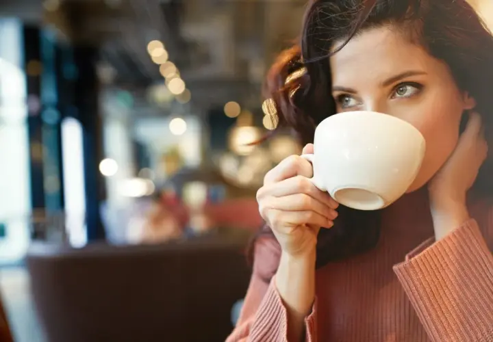 En kvinna med mörkt hår dricker kaffe ur en vit kopp. Hon har en stickad tröja på sig och sitter på ett café. Kanske är det en morgon med fastande mage kaffe.