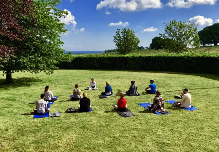 En grupp människor sitter på yogamattor på en gräsmatta under en klarblå himmel. En instruktör leder dem i en övning under ett yoga weekend.
