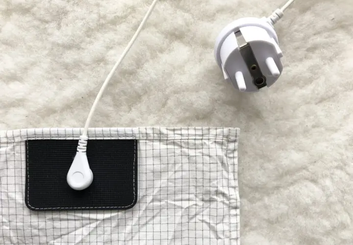 En vit jordad kabel med kontakt ligger på en vit, fluffig yta. En del av en rutig duk med en svart ficka syns. Lavasten egenskaper kan vara relaterade till jordning.