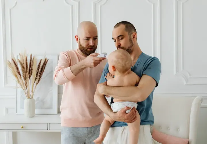 Två pappor kollar febern på sitt barn. En pappa håller barnet, den andra mäter temperaturen. Frågan "hur får man bort snuva" kan dyka upp när man tar hand om små barn.
