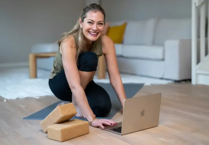 Glad kvinna förbereder sig för en lugnande restorative yoga-session med hjälp av en laptop och yogablock.
