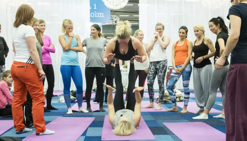 Grupp kvinnor deltar i en yogaklass. En kvinna lyfter en annan i en partnerövning, vilket visar den fysiska och andliga aspekten av yoga, liknande en andlig religion.