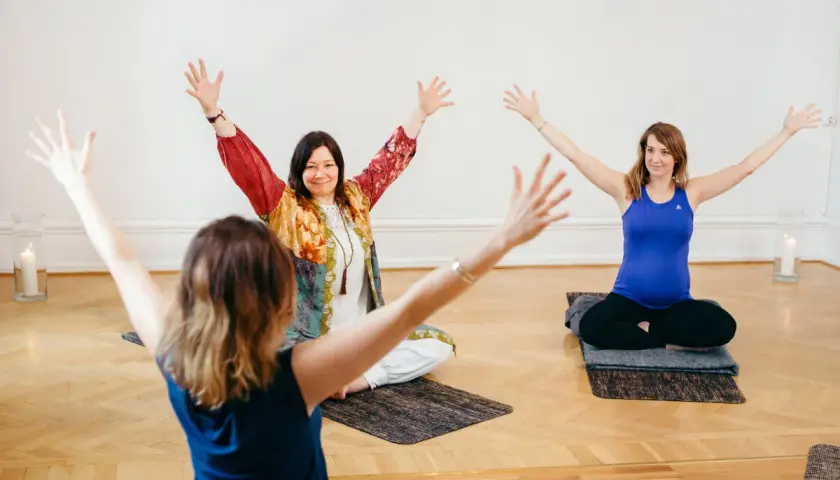 Tre kvinnor sitter på yogamattor med armarna uppåt. En av dem är gravid. En inspirerande yoga utbildning distans.