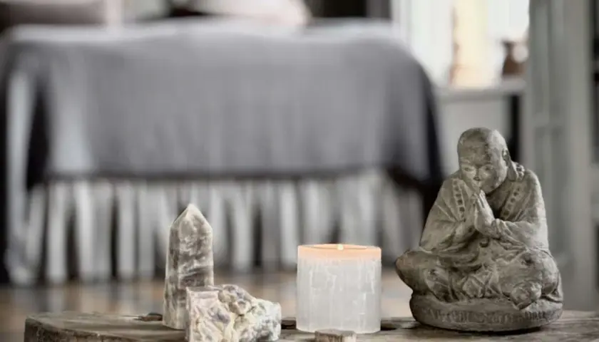 En stilla Buddha-statyett, en tänd ljuslykta och stenar, kanske med honungskalcit egenskaper, på ett träbord framför en säng.