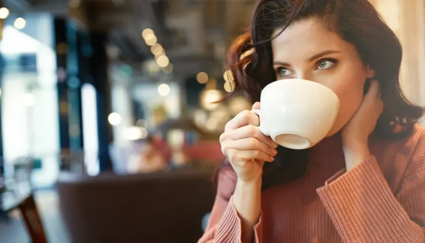 En kvinna med mörkt hår dricker kaffe ur en vit kopp. Hon har en stickad tröja på sig och sitter på ett café. Kanske är det en morgon med fastande mage kaffe.