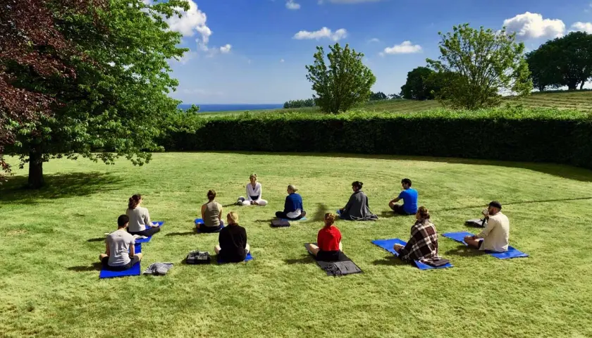 En grupp människor sitter på yogamattor på en gräsmatta under en klarblå himmel. En instruktör leder dem i en övning under ett yoga weekend.