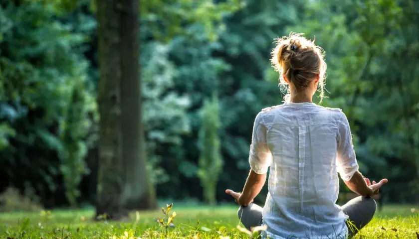 En person sitter i lotusställning i en grönskande park, med ryggen mot kameran. Solen lyser genom träden och skapar en varm glöd. En stund av lugn och eye gazing.