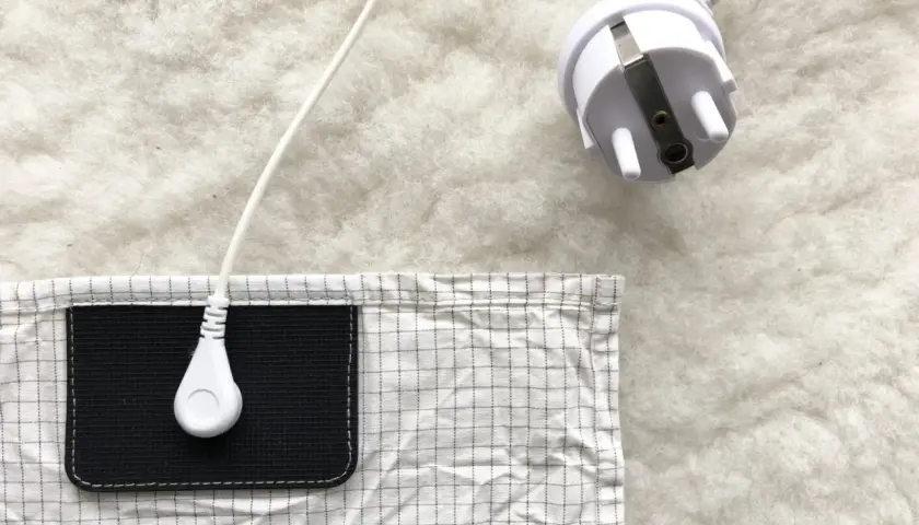 En vit jordad kabel med kontakt ligger på en vit, fluffig yta. En del av en rutig duk med en svart ficka syns. Lavasten egenskaper kan vara relaterade till jordning.