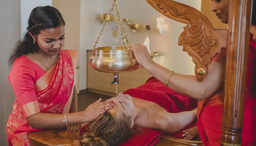 Två kvinnor i sari utför en indisk massage med olja som rinner ner över en persons ansikte.