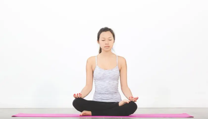 En kvinna sitter i lotusställning på en rosa yogamatta, utövar pranayama yoga med slutna ögon.