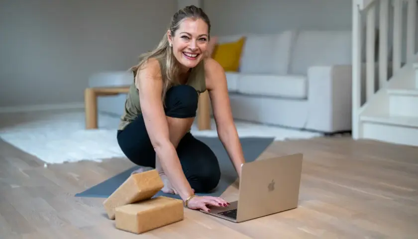 Glad kvinna förbereder sig för en lugnande restorative yoga-session med hjälp av en laptop och yogablock.