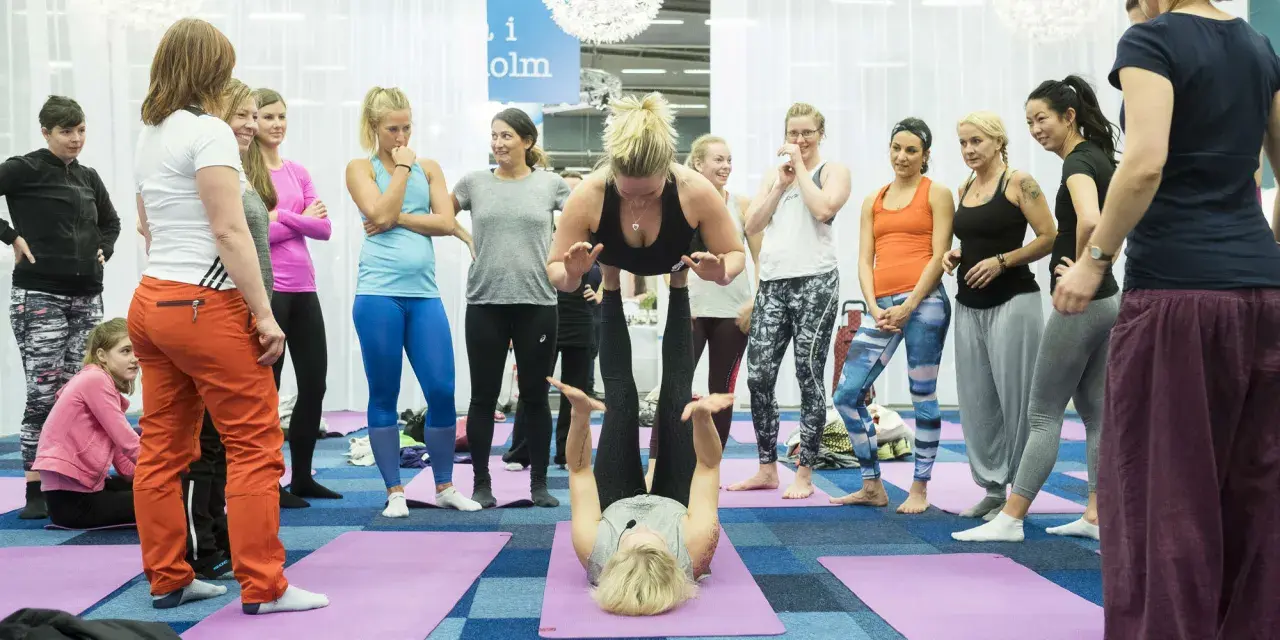 Grupp kvinnor deltar i en yogaklass. En kvinna lyfter en annan i en partnerövning, vilket visar den fysiska och andliga aspekten av yoga, liknande en andlig religion.