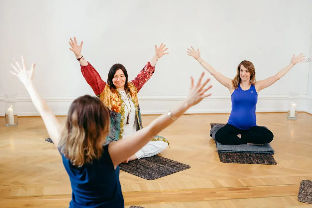 Tre kvinnor sitter på yogamattor med armarna uppåt. En av dem är gravid. En inspirerande yoga utbildning distans.