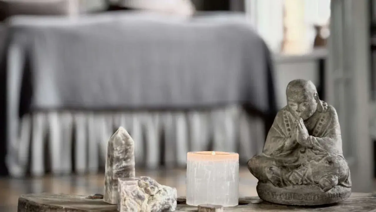 En stilla Buddha-statyett, en tänd ljuslykta och stenar, kanske med honungskalcit egenskaper, på ett träbord framför en säng.