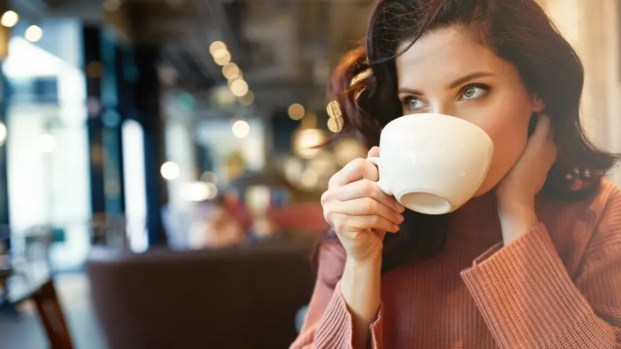 En kvinna med mörkt hår dricker kaffe ur en vit kopp. Hon har en stickad tröja på sig och sitter på ett café. Kanske är det en morgon med fastande mage kaffe.