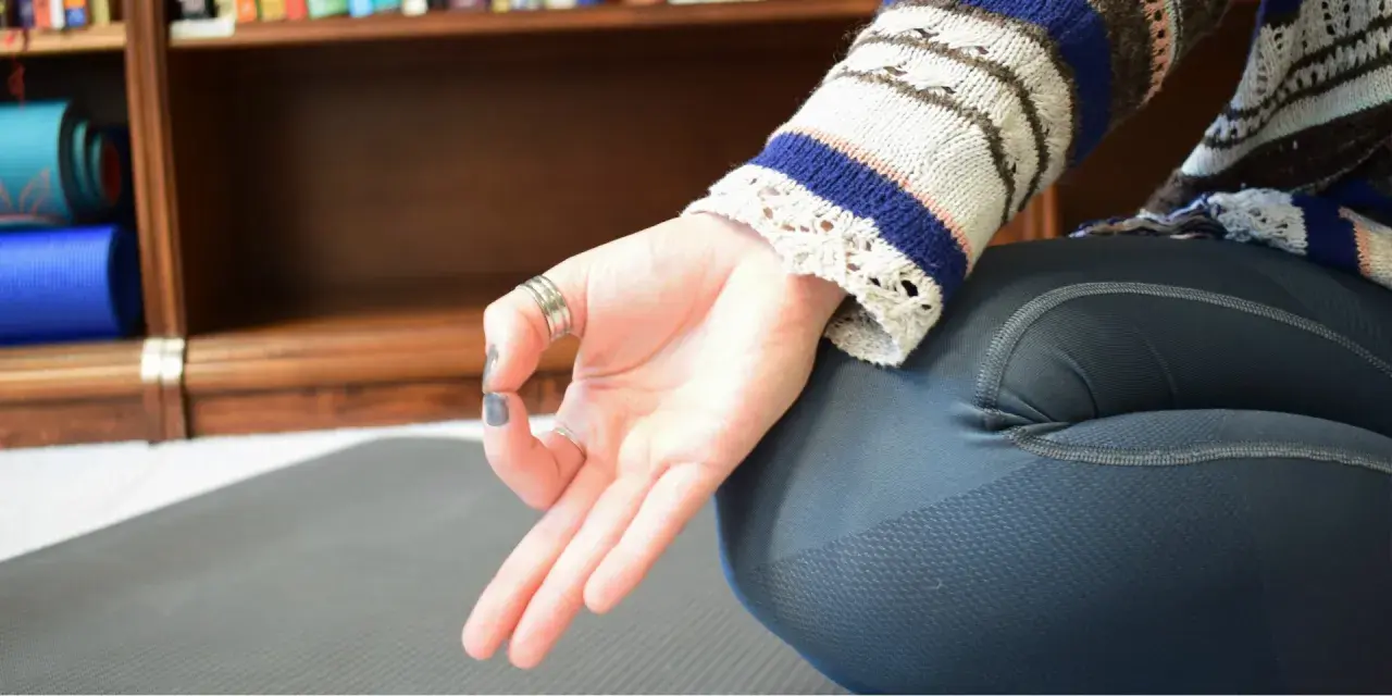 En hand i anjali mudra vilar på ett knä. Bakom syns en bokhylla med yogamattor.