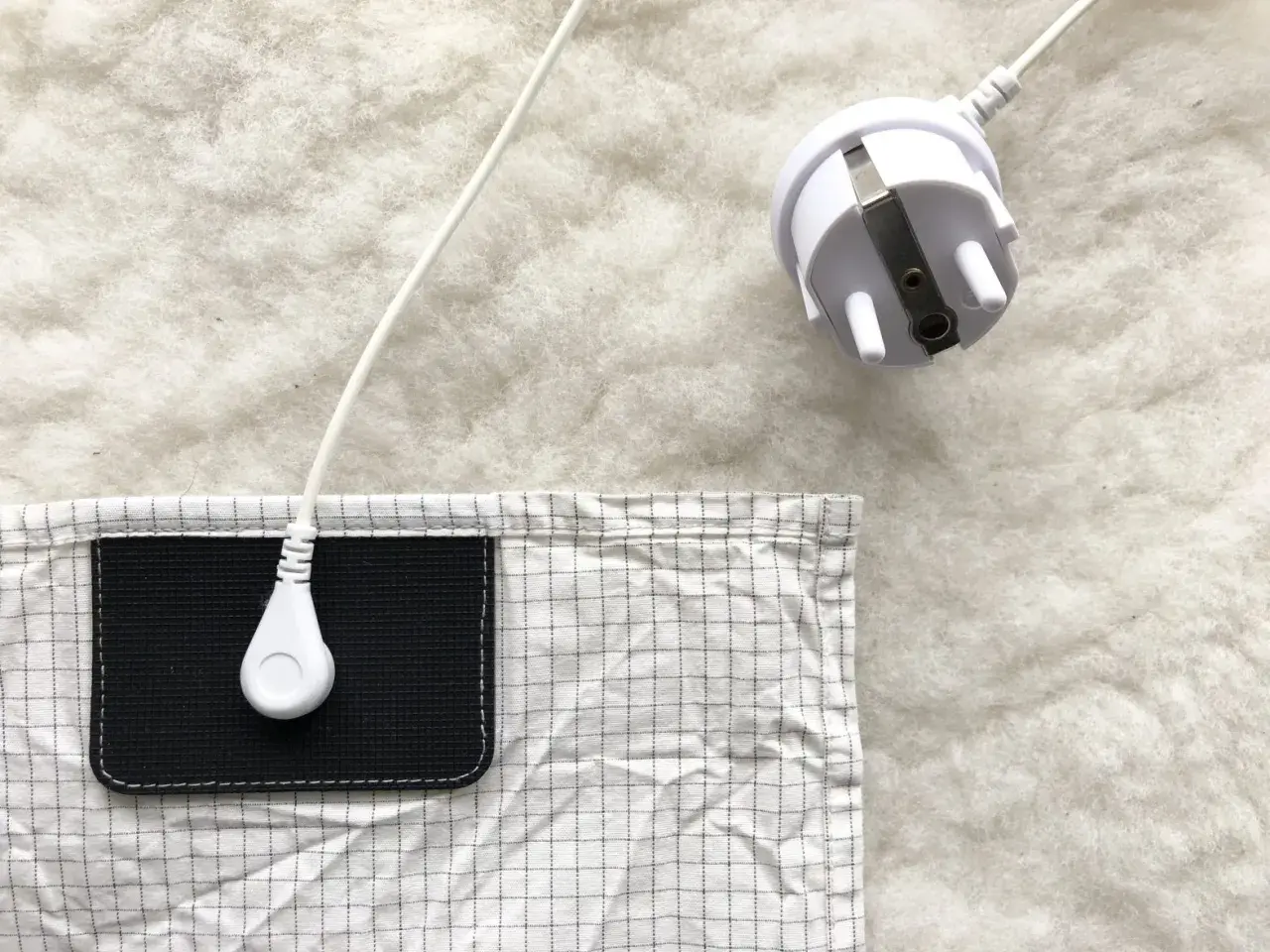 En vit jordad kabel med kontakt ligger på en vit, fluffig yta. En del av en rutig duk med en svart ficka syns. Lavasten egenskaper kan vara relaterade till jordning.