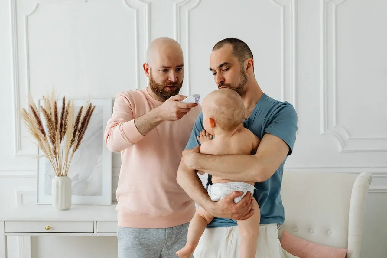 Två pappor kollar febern på sitt barn. En pappa håller barnet, den andra mäter temperaturen. Frågan "hur får man bort snuva" kan dyka upp när man tar hand om små barn.