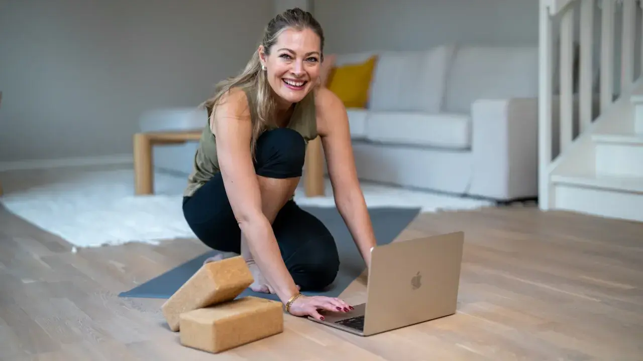 Glad kvinna förbereder sig för en lugnande restorative yoga-session med hjälp av en laptop och yogablock.