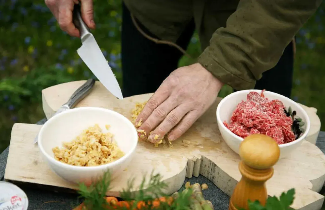 Förbereder eko mat utomhus. En hand hackar ingredienser på en träskärbräda bredvid skålar med köttfärs och en annan ingrediens.
