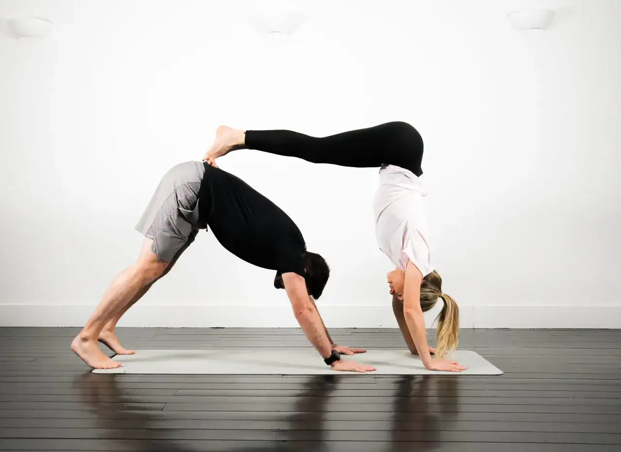 Tv&aring; personer utf&ouml;r avancerad yoga. Mannen &auml;r i ned&aring;tg&aring;ende hund, kvinnan balanserar p&aring; hans rygg i en handst&aring;ende.