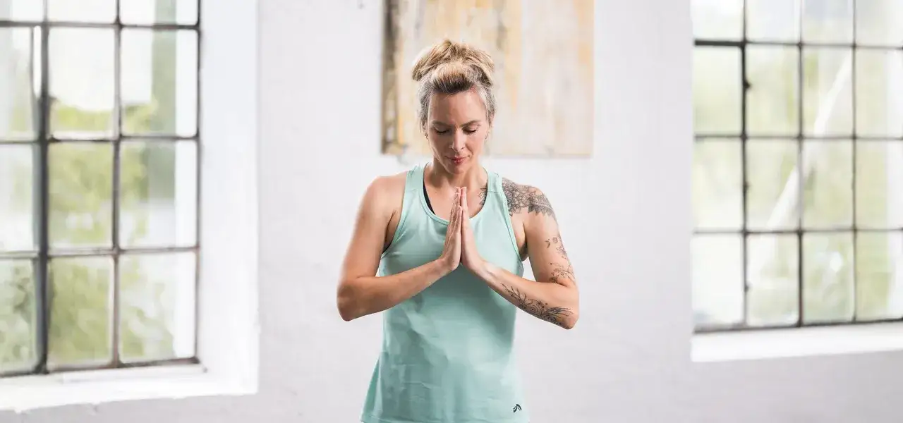 Kvinna i ljusgr&ouml;n topp med h&auml;nderna i b&ouml;nest&auml;llning, redo f&ouml;r yoga. En lugn stund med en kopp yoga te v&auml;ntar.