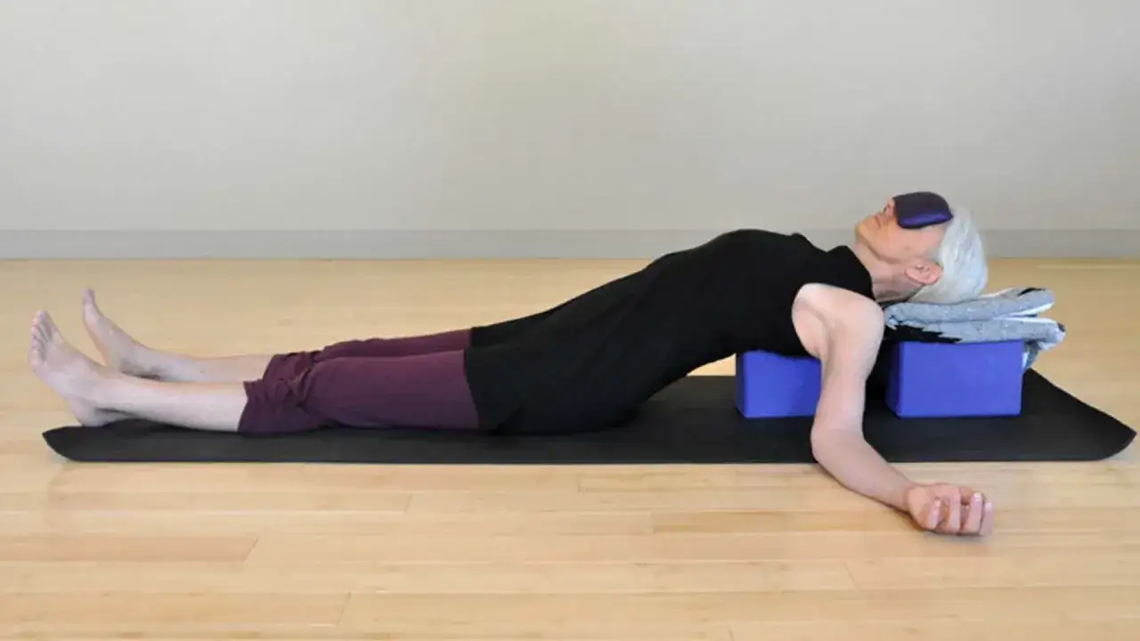 &Auml;ldre kvinna vilar p&aring; rygg p&aring; yogamatta med &ouml;gonbindel. Yoga f&ouml;r rygg och nacke med st&ouml;d av block och filtar.