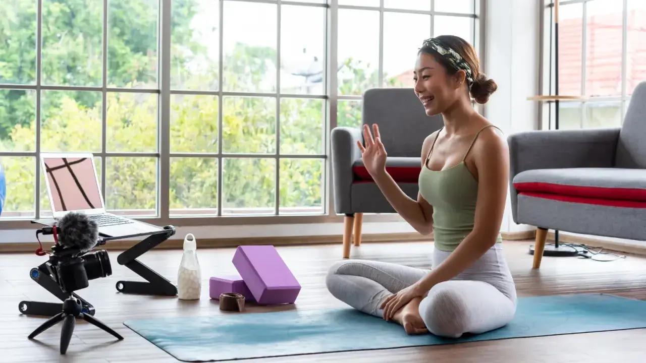 Kvinna vinkar till kameran, redo f&ouml;r yoga utbildning distans. Utrustning f&ouml;r inspelning och yogablock syns.