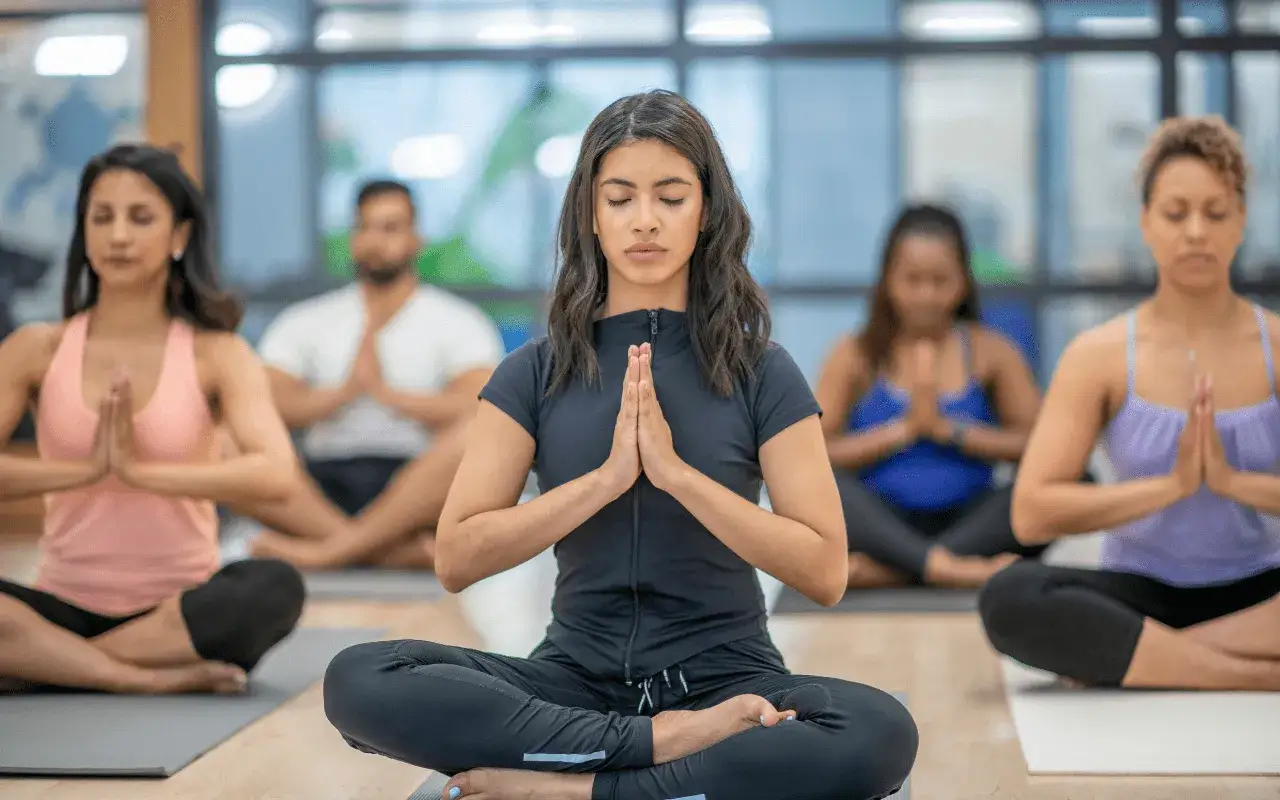 En grupp m&auml;nniskor utf&ouml;r yoga poses i en studio. De sitter p&aring; mattor med slutna &ouml;gon och h&auml;nderna i b&ouml;neposition.