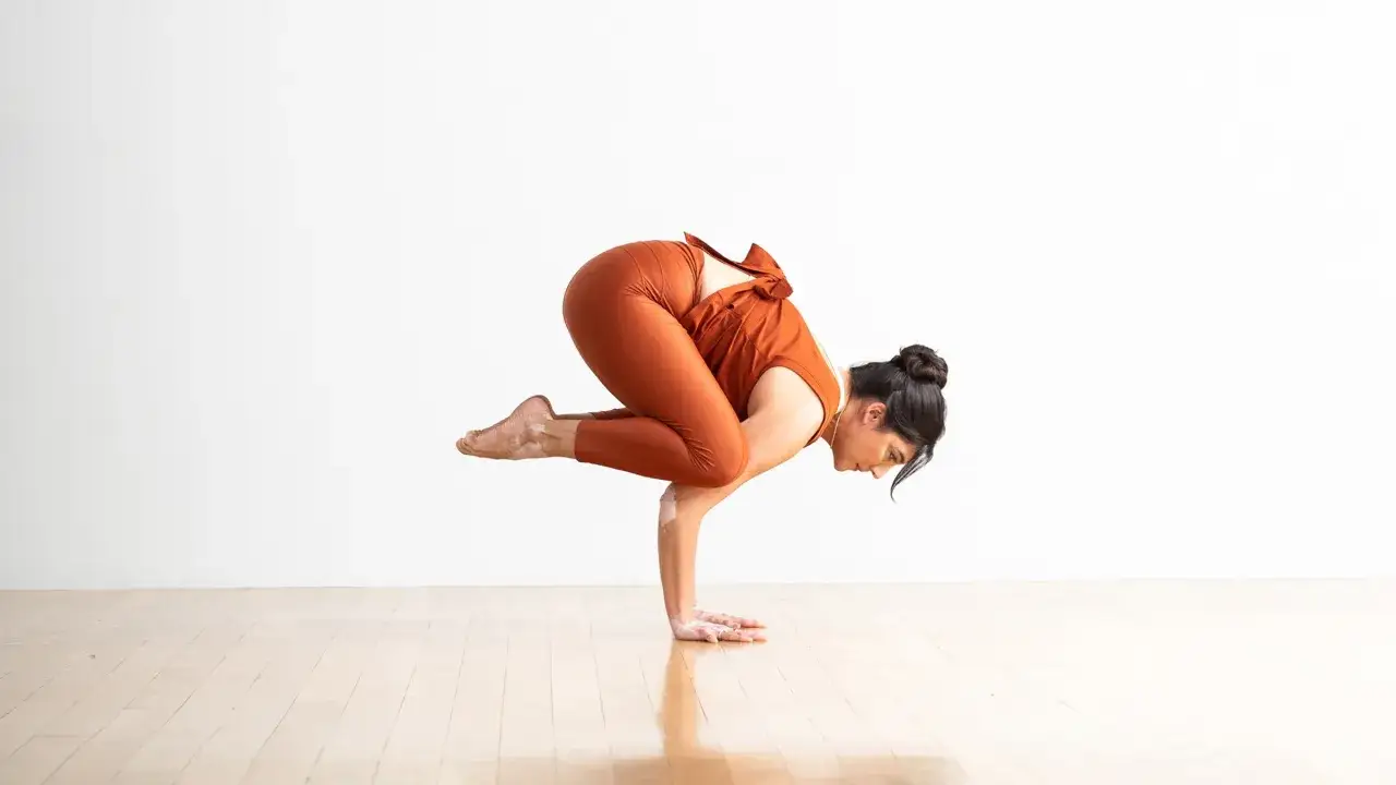 En kvinna utf&ouml;r kr&aring;kan yoga, en utmanande balansst&auml;llning, med en orange outfit mot en vit bakgrund.