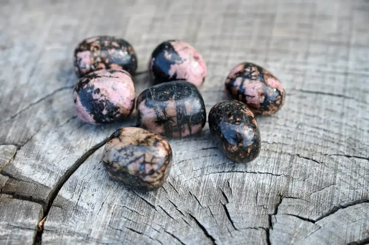 Sju polerade stenar med rosa och svarta m&ouml;nster ligger p&aring; en tr&auml;yta. Dessa stenar, k&auml;nda f&ouml;r sina helande rodonit egenskaper, ser ut att vara redo att anv&auml;ndas i smycken eller meditation.