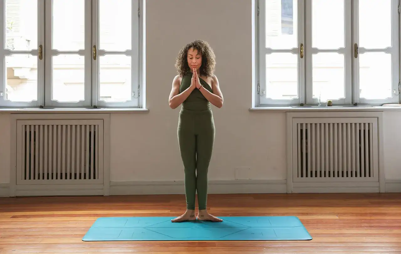 En kvinna i gröna träningskläder står på en yogamatta med händerna i böneposition framför bröstet. Hon utstrålar lugn och säger 