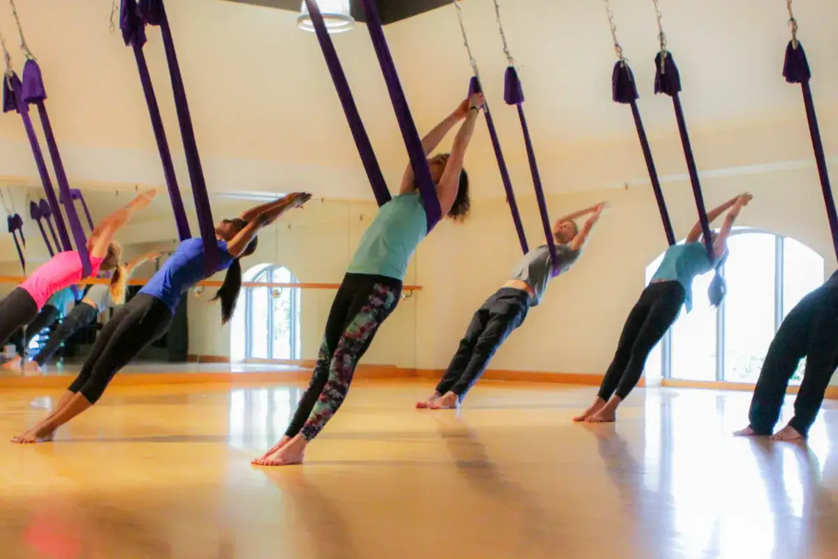 M&auml;nniskor ut&ouml;var flying yoga i lila tyger. De str&auml;cker sig bak&aring;t med armarna upp&aring;t.