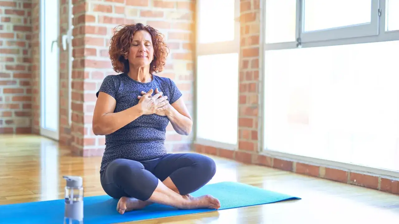 Kvinna med slutna &ouml;gon sitter i lotusst&auml;llning p&aring; en yogamatta, med h&auml;nderna &ouml;ver hj&auml;rtat. Hon ut&ouml;var yoga mot &aring;ngest och finner lugn.
